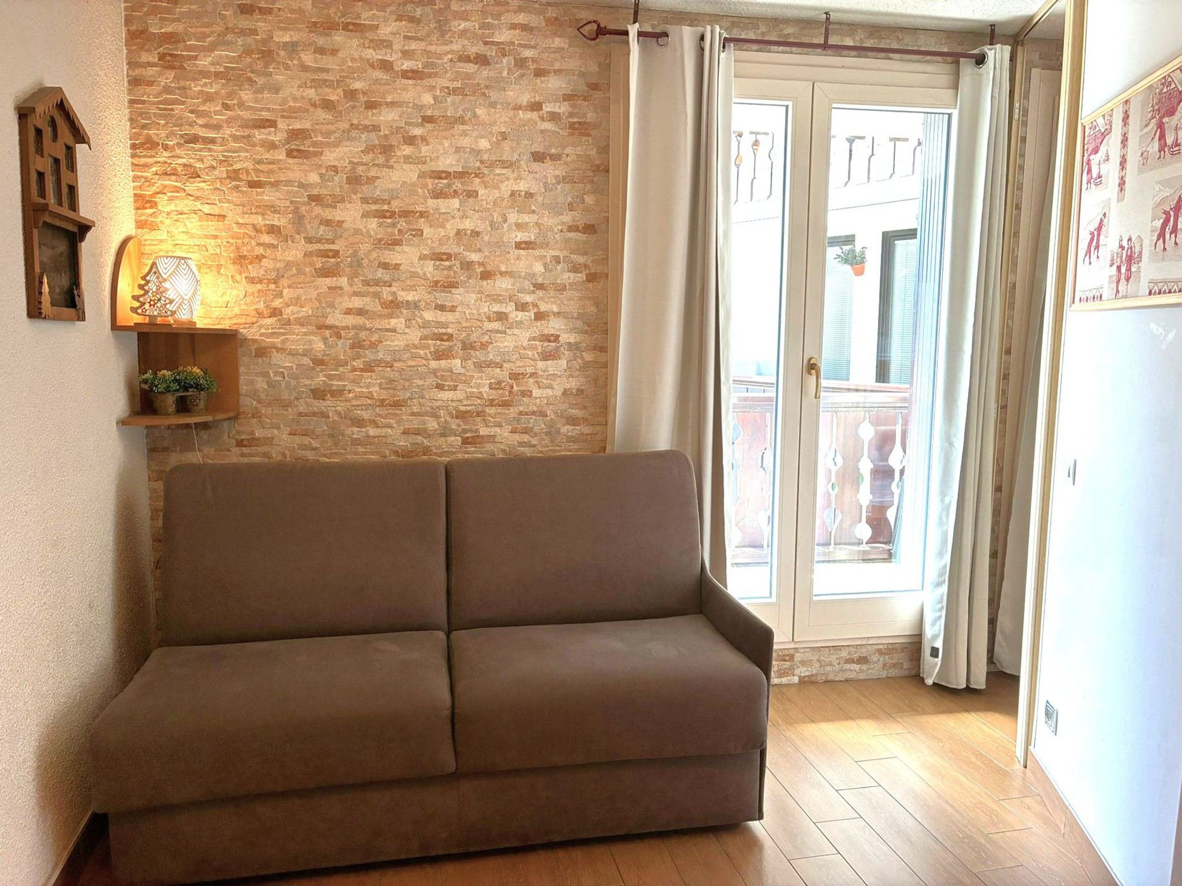 A room with a brown sofa, a brick wall, a wooden floor, and a sliding glass door leading to an outdoor balcony.