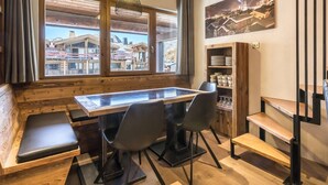 A cozy dining area with a wooden table, chairs, and a bench. Large windows offer a view of a mountain village. - Luxury Apartment with WiFi and Parking, Ski-in/Ski-out, Town Center (Val-d'Isère)