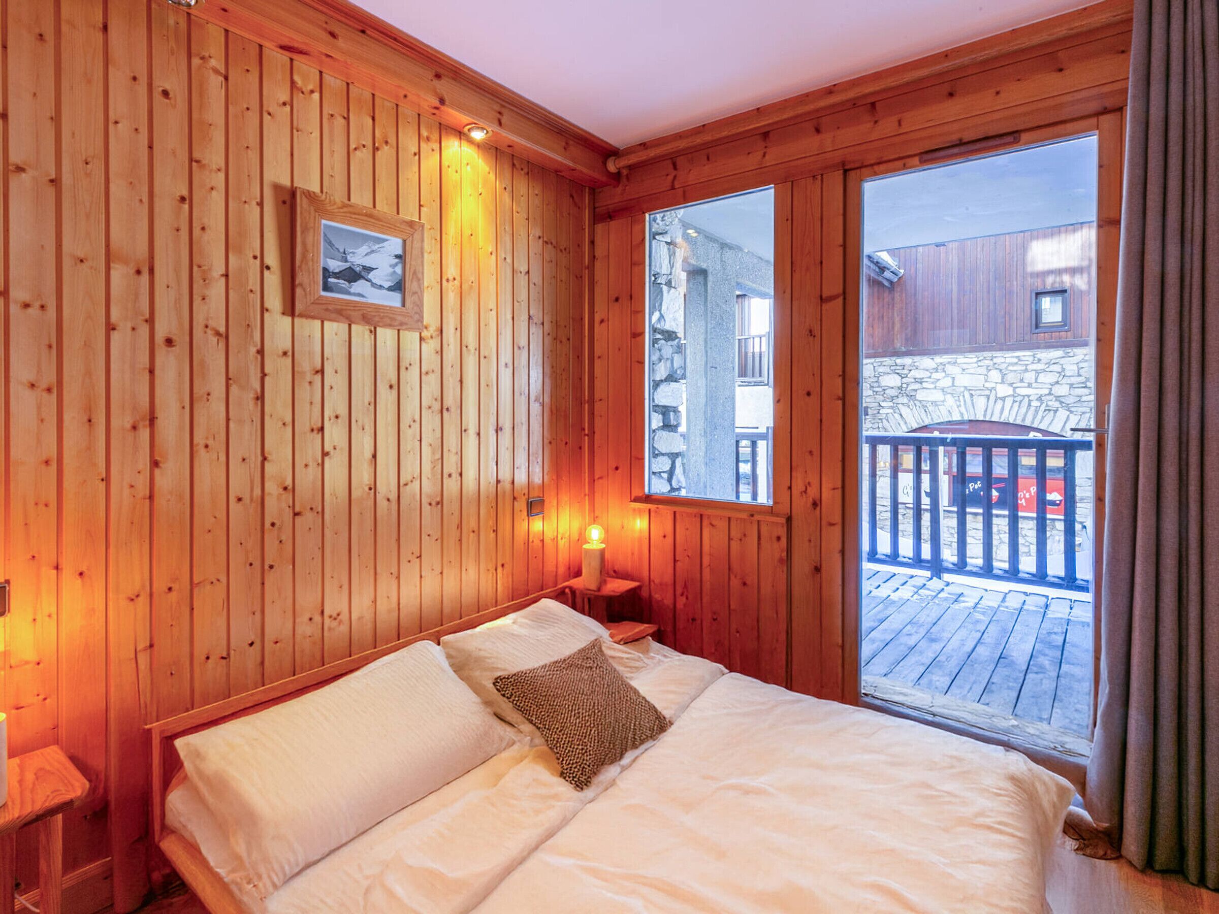 A wooden room with a bed, a picture on the wall, and a window with a view of a balcony.