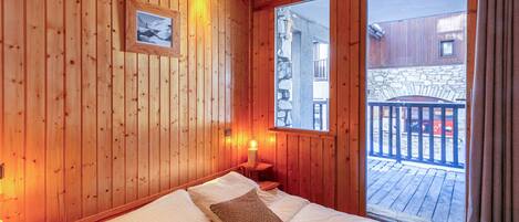 A wooden room with a bed, a picture on the wall, and a window with a view of a balcony.