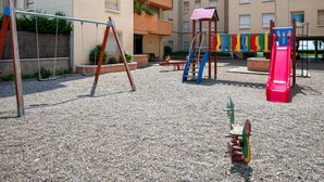 An outdoor playground with swings, a slide, and climbing structures, surrounded by a gravel area and a multi-story building in the background. - Seaside Apartment with Wi-Fi and Parking - Torredembarra (Torredembarra)