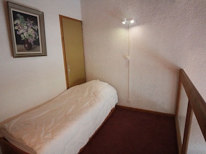 A small, single-bed room with a wooden headboard, a door, and a wall-mounted light fixture.