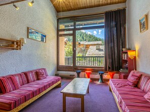 Ein gemütliches Zimmer mit Holzbalkendecke, einem großen Fenster mit Blick auf einen Berg und Gebäude, einer Couch, einem Tisch und einem Bücherregal mit Büchern und einem Korb.