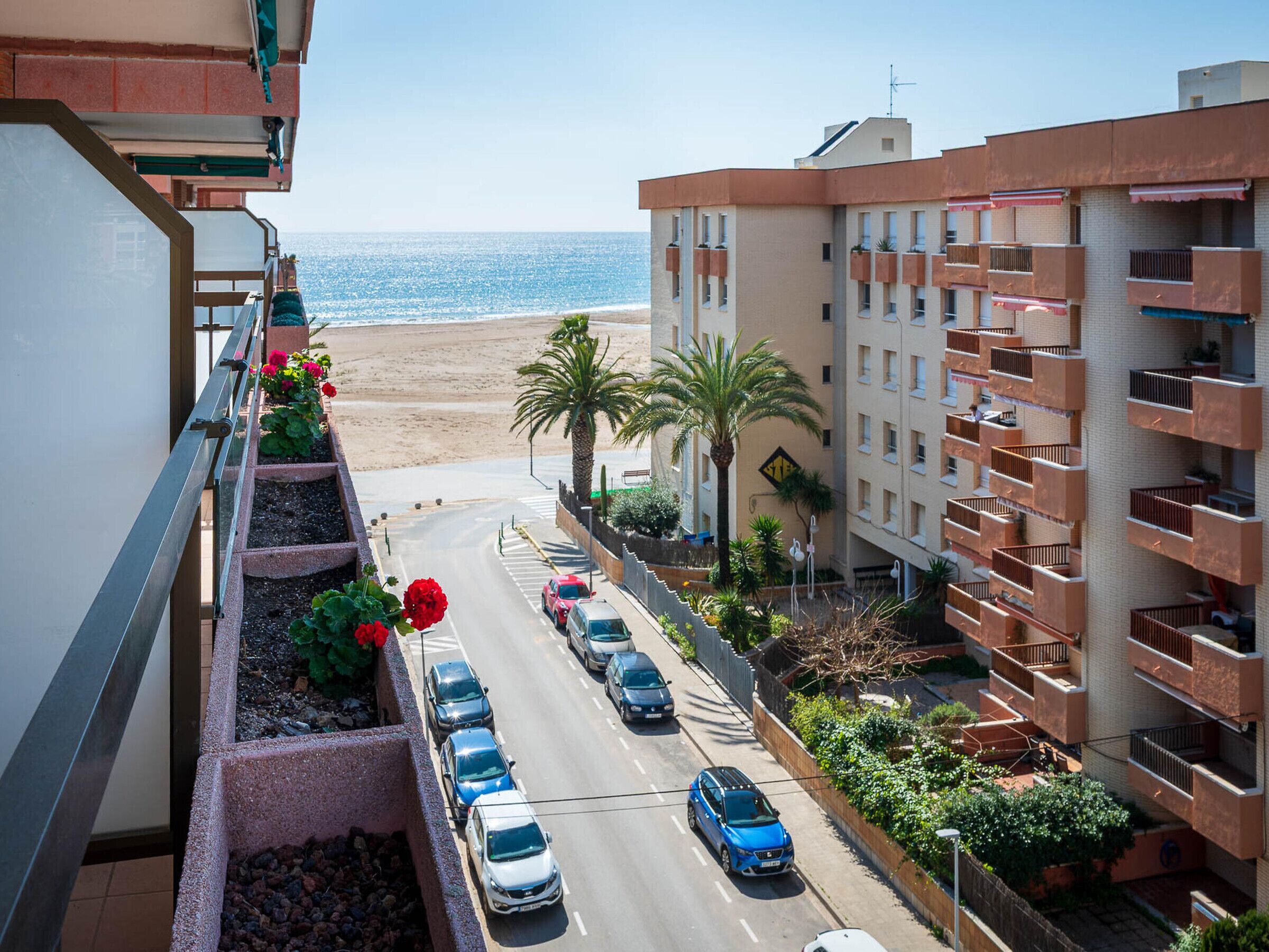 Un bâtiment situé sur la côte, doté de nombreux balcons qui donnent sur une plage et une route bordée de voitures garées.
