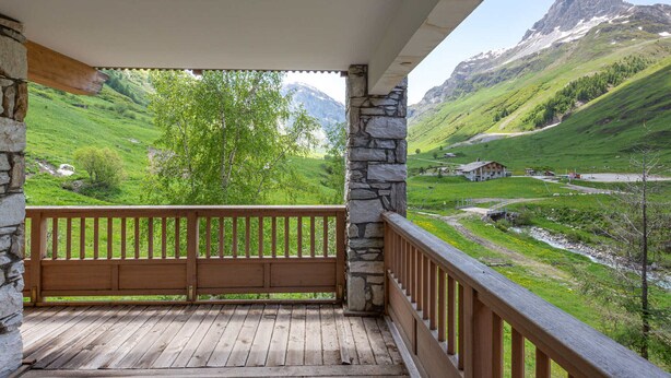 Un balcon en bois, soutenu par des piliers en pierre, qui offre une vue sur une vallée verdoyante et des montagnes.