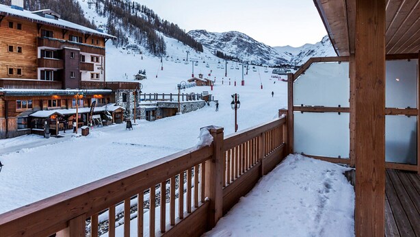 A wooden balcony with a view of a snow-covered ski slope and buildings in the background.