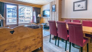 A wooden room with a bed, a dining table with red chairs, a TV on the wall, and a window with a view of buildings.