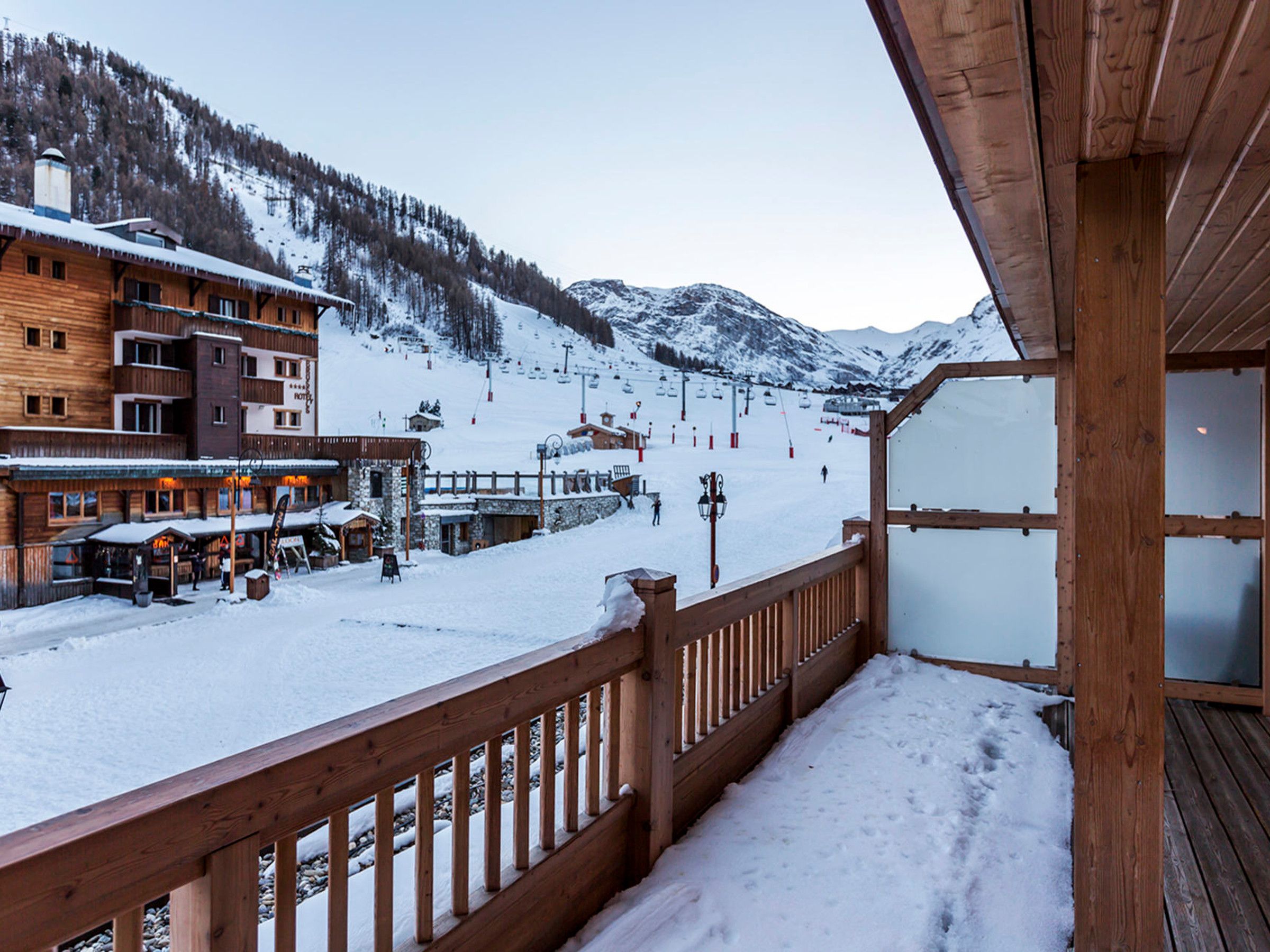 A wooden balcony with a view of a snow-covered ski slope and buildings in the background.