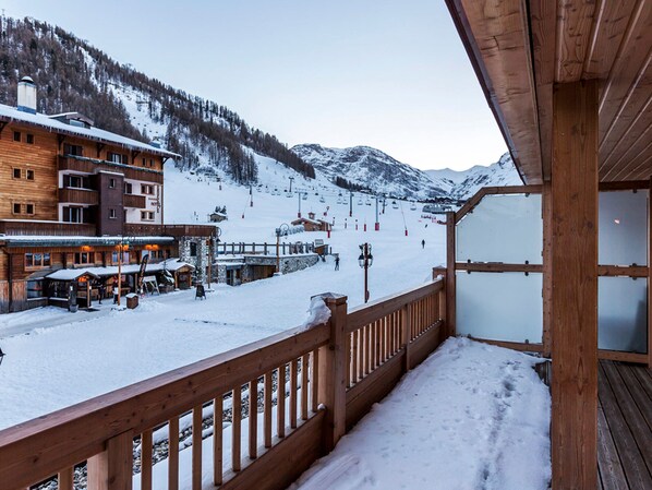 A wooden balcony with a view of a snow-covered ski slope and buildings in the background.