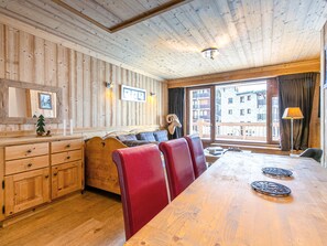 A wooden interior with a long dining table, red upholstered chairs, a wooden cabinet, and a large window offering a view of buildings outside. - Elegant Apartment with Terrace, WiFi, Ski-In/Ski-Out in Val-d'Isère (Val-d'Isère)