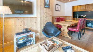 A cozy room with a wooden dining table, red chairs, a black chair, a TV, and a kitchen area with wooden cabinets.