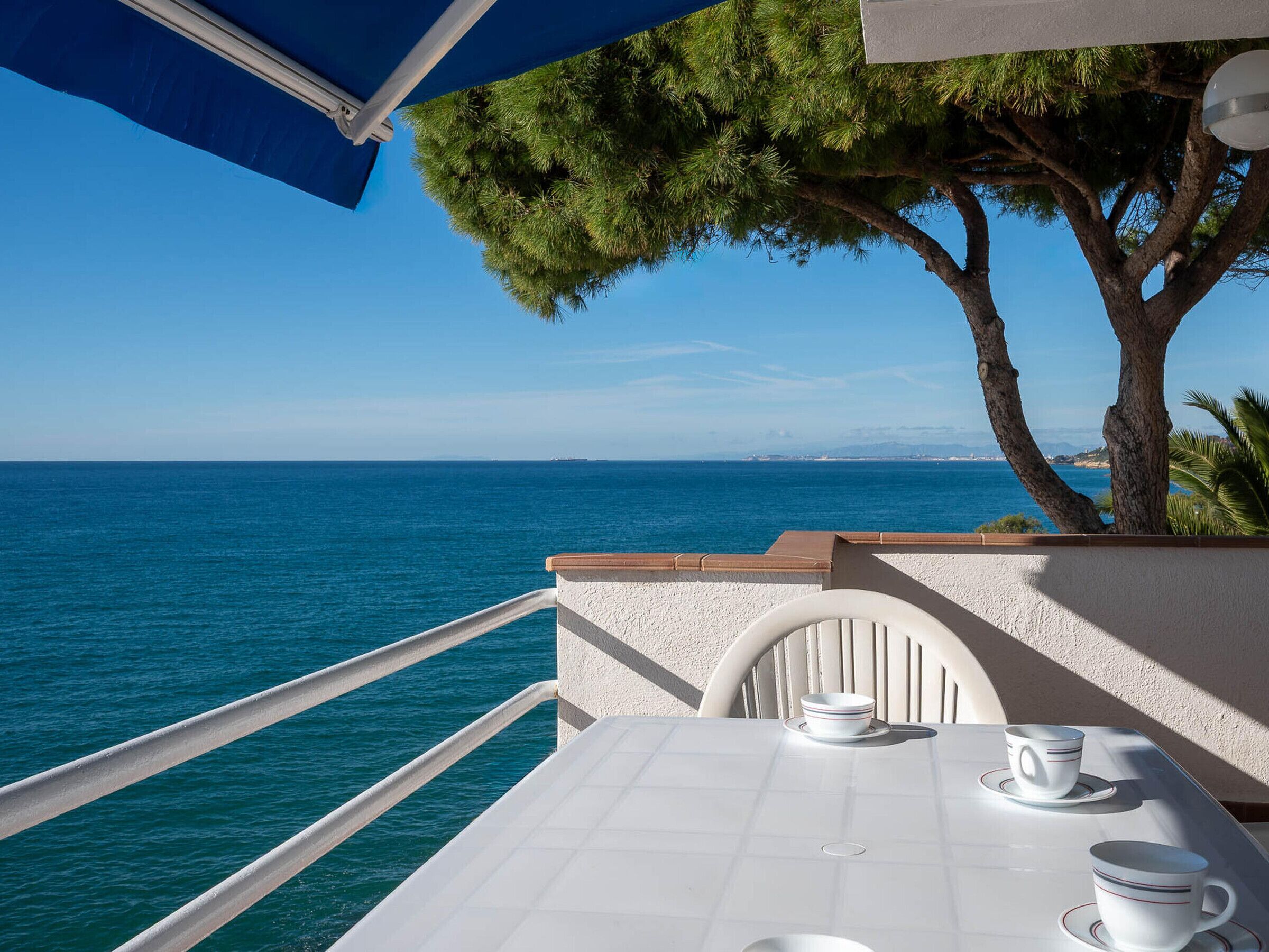 A balcony with a table set for two, overlooking a clear blue sea and a distant coastline.