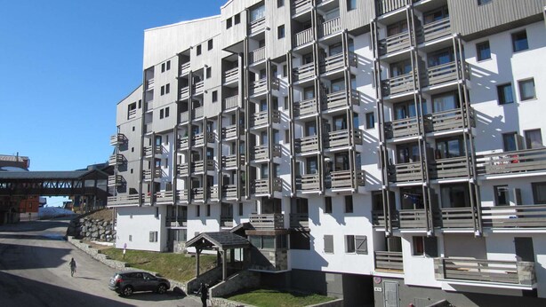 Un edificio residencial de varios pisos con balcones, un acceso cubierto y una zona de estacionamiento.