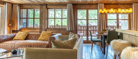 A rustic cabin interior with a wooden ceiling, leather sofas, a dining area, and large windows.