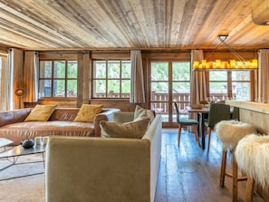 A rustic cabin interior with a wooden ceiling, leather sofas, a dining area, and large windows.