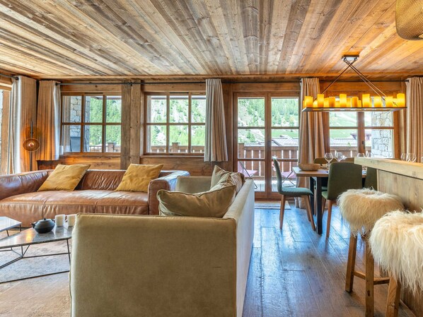 A rustic cabin interior with a wooden ceiling, leather sofas, a dining area, and large windows.