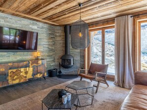 Ein gemütliches Wohnzimmer mit Steinwand, Kamin, Fernseher und Blick ins Freie.