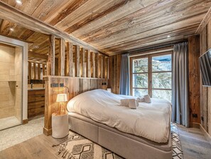 A bedroom with a wooden ceiling, a bed with white linens, a bedside table with a lamp, and a window with curtains.