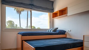 Una habitación con una cama litera, un ventanal con vistas a árboles, un estante de madera pequeño y una pared blanca.