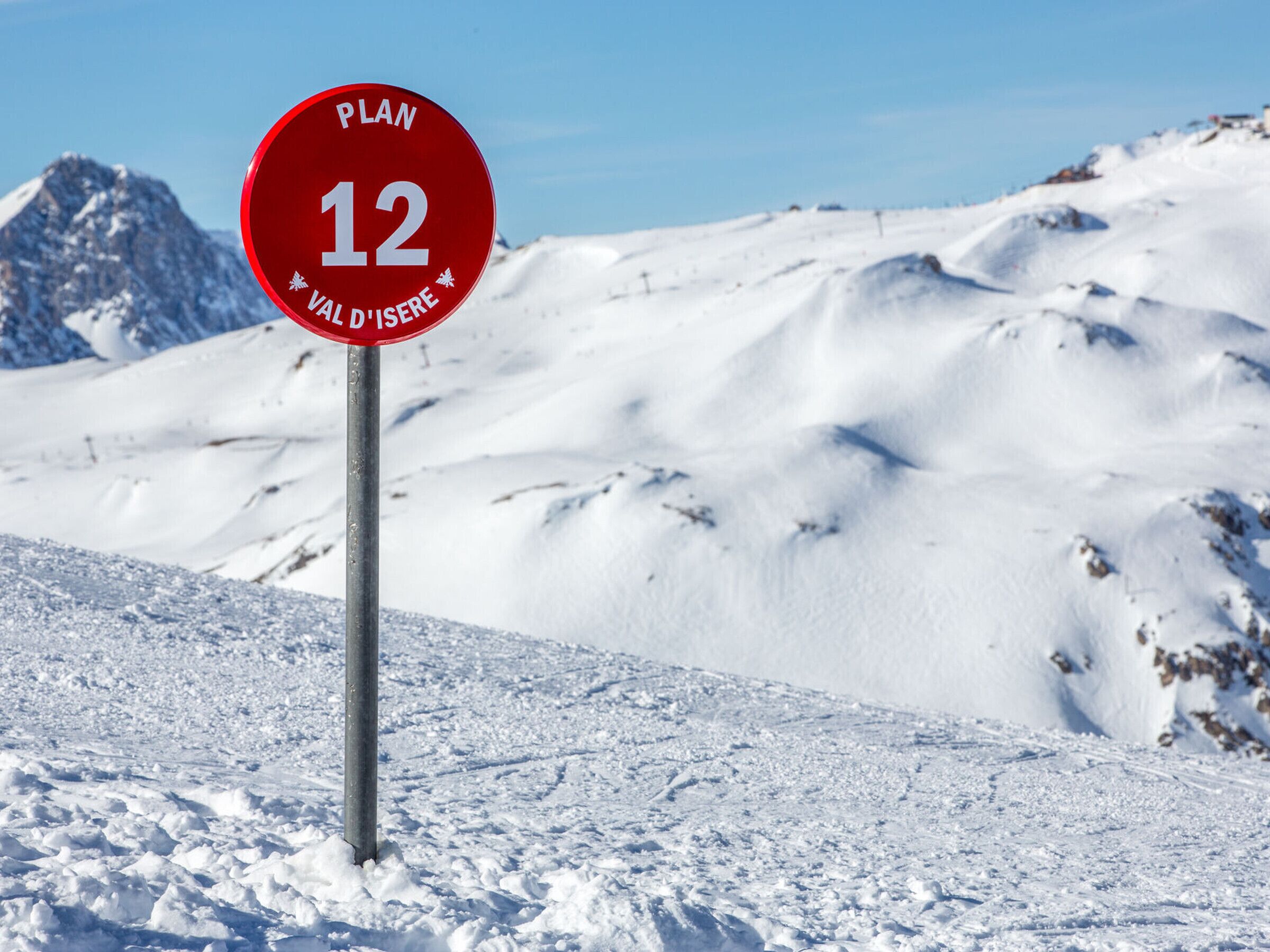 Um sinal vermelho com o número 12 e o texto 'VAL D'ISERE' ao fundo de uma montanha coberta de neve.