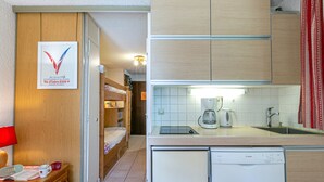 Ett kompakt hotellrum med ett litet pentry, en säng, ett litet bord med en lampa och ett inramat certifikat på väggen.