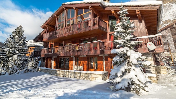 A multi-story wooden chalet with balconies, surrounded by snow-covered trees and a clear blue sky.