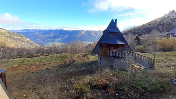 A small wooden cabin with a steeply pitched roof, surrounded by a grassy landscape and hills in the background.