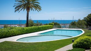 A swimming pool with a curved edge, surrounded by well-maintained grass and hedges, with a view of the sea and boats in the background.