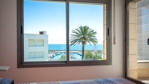 A bedroom with a large window overlooking a marina and the sea, a bed with a patterned quilt, and a door to the right.