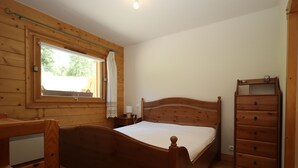 Ein Schlafzimmer mit Holzrahmen, einem Bett, einer Kommode, einem Sessel und einem Fenster mit Vorhang.