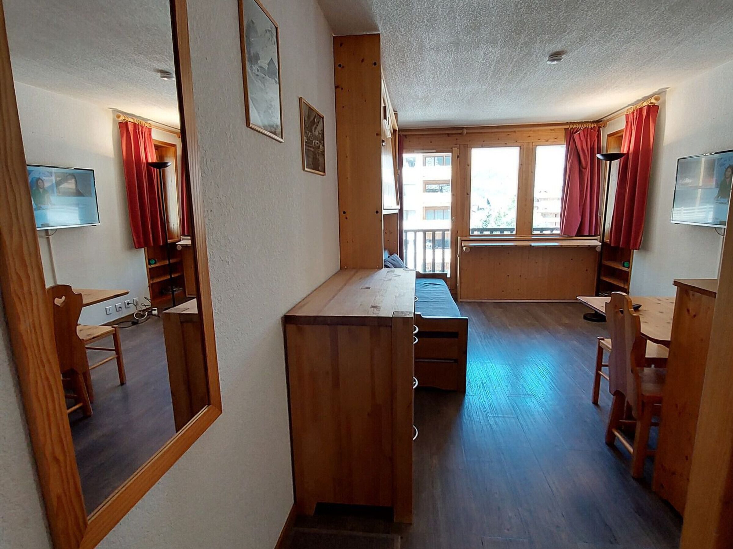 A compact room with a wooden desk, a bed, and a small table. There are two windows with red curtains, and a mirror on the left wall.