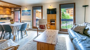 Een gezellige woonkamer met een houten tafel, een bank en een televisie die aan een houten wand is bevestigd. Er is een eethoek met stoelen en een tafel, en een keuken met houten kasten.