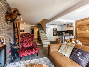 Ein modernes Wohnzimmer mit Kamin, einem roten, gemusterten Sessel, einer braunen Ledersofa und einer Küche im Hintergrund.