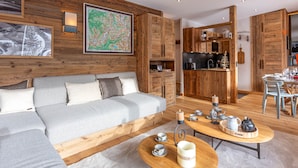 A modern living room with a sofa, coffee table, and a kitchen area with wooden cabinets and a dining table. - Luxury Chalet Apartment, Ski-In/Ski-Out, Val-d’Isère (Val-d'Isère)
