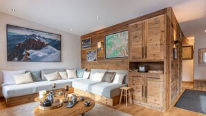 A modern living room with a wooden accent wall, a sectional sofa, a coffee table, and a small stool. - Luxury Chalet Apartment, Ski-In/Ski-Out, Val-d’Isère (Val-d'Isère)