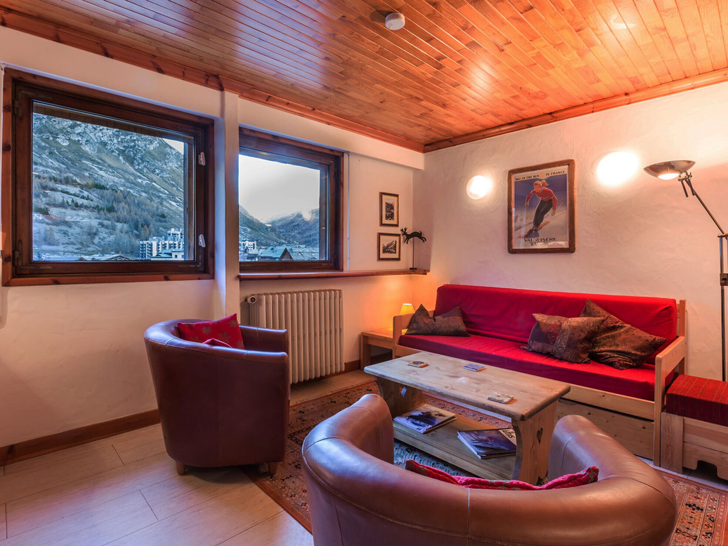 Ein gemütliches Wohnzimmer mit einem roten Sofa, Ledersesseln und einem Holztisch. Große Fenster bieten Blick auf verschneite Berge.