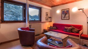 Uma sala de estar acolhedora com um sofá vermelho, poltronas de couro e uma mesa de centro de madeira. Janelas amplas oferecem vista para montanhas nevadas.