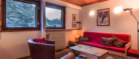 Un salon chaleureux avec un canapé rouge, des fauteuils en cuir et une table basse en bois. De grandes fenêtres offrent une vue sur des montagnes enneigées.