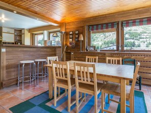 A wooden interior with a dining table, chairs, bar stools, and a kitchen area with a window overlooking a mountainous landscape. - Val-d'Isère: Spacious apartment with balcony, sleeps 7 (Val-d'Isère)