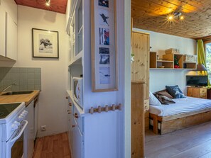 A compact kitchen with a sink, stove, and wooden cabinets. A bed with a TV and storage shelves is visible in the adjacent room. - Comfortable studio in Val-d'Isère center with Wifi, pets accepted (Val-d'Isère)