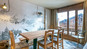 Un comedor con mesa y sillas de madera, un mural con paisaje nevado de montaña y vista a un edificio de madera a través de un amplio ventanal.