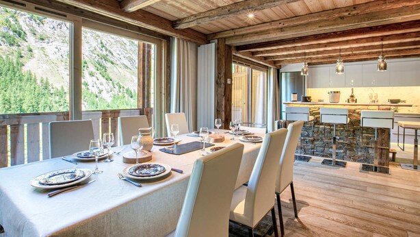 Un espace repas avec une longue table dressée pour six personnes, des poutres en bois au plafond et une cuisine en arrière-plan.