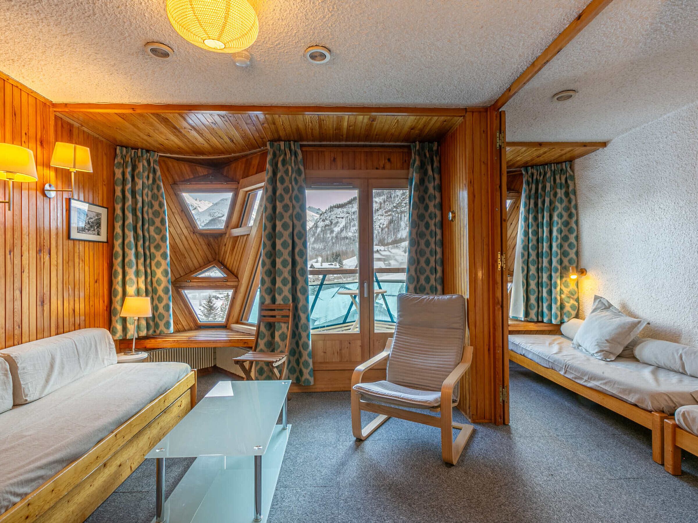 Un chalet confortable aux murs en bois, avec un canapé, un fauteuil et une table en verre. De grandes fenêtres offrent une vue imprenable sur les montagnes.