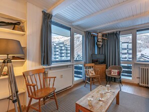 A cozy room with a wooden table, chairs, a lamp, a television, and large windows offering a view of a snowy mountain.