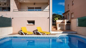 Un área de piscina con dos sillas de sol amarillas, una piscina azul cristalina y un edificio con balcón y una ventana pequeña.