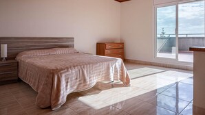Un dormitorio con cama, mesita de noche, cómoda y un amplio ventanal que ofrece vistas del cielo.