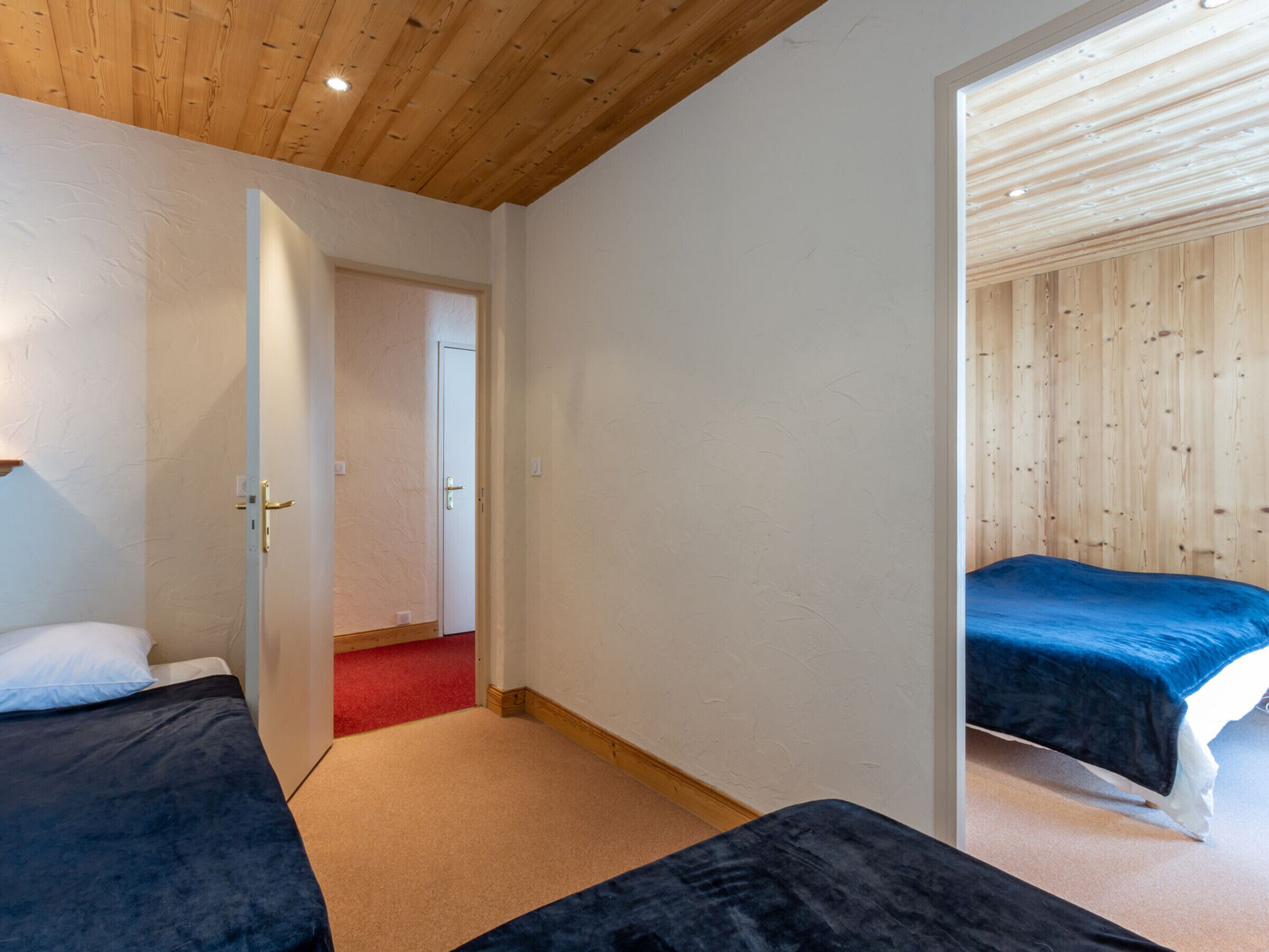 A bedroom with two beds, wooden ceiling, and a door leading to another room.