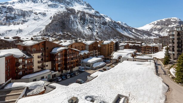 Un centro turístico de montaña cubierto de nieve con varios edificios de alojamiento, un telesilla y un cielo azul despejado.