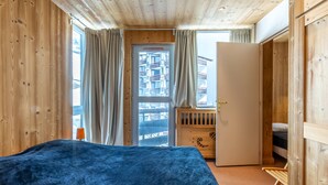Un dormitorio con paredes de madera, una cama, un ventanal con cortinas y una puerta que da a otra habitación.