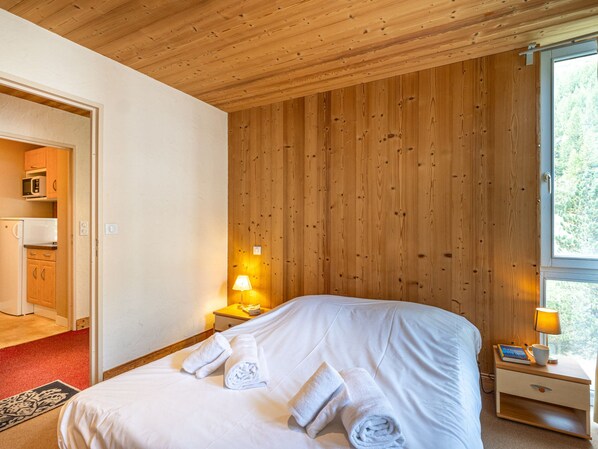 A hotel room with a wooden ceiling, a bed with white linens, a nightstand with a lamp, and a view of the outdoors through a window.